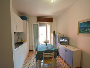 A compact kitchen with a dining table, chairs, a TV, and a painting on the wall. - Central Apartment Near the Beach With Parking (Lignano Sabbiadoro)