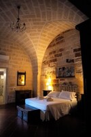 A bedroom with a stone wall, a bed, a nightstand, a lamp, and a painting.