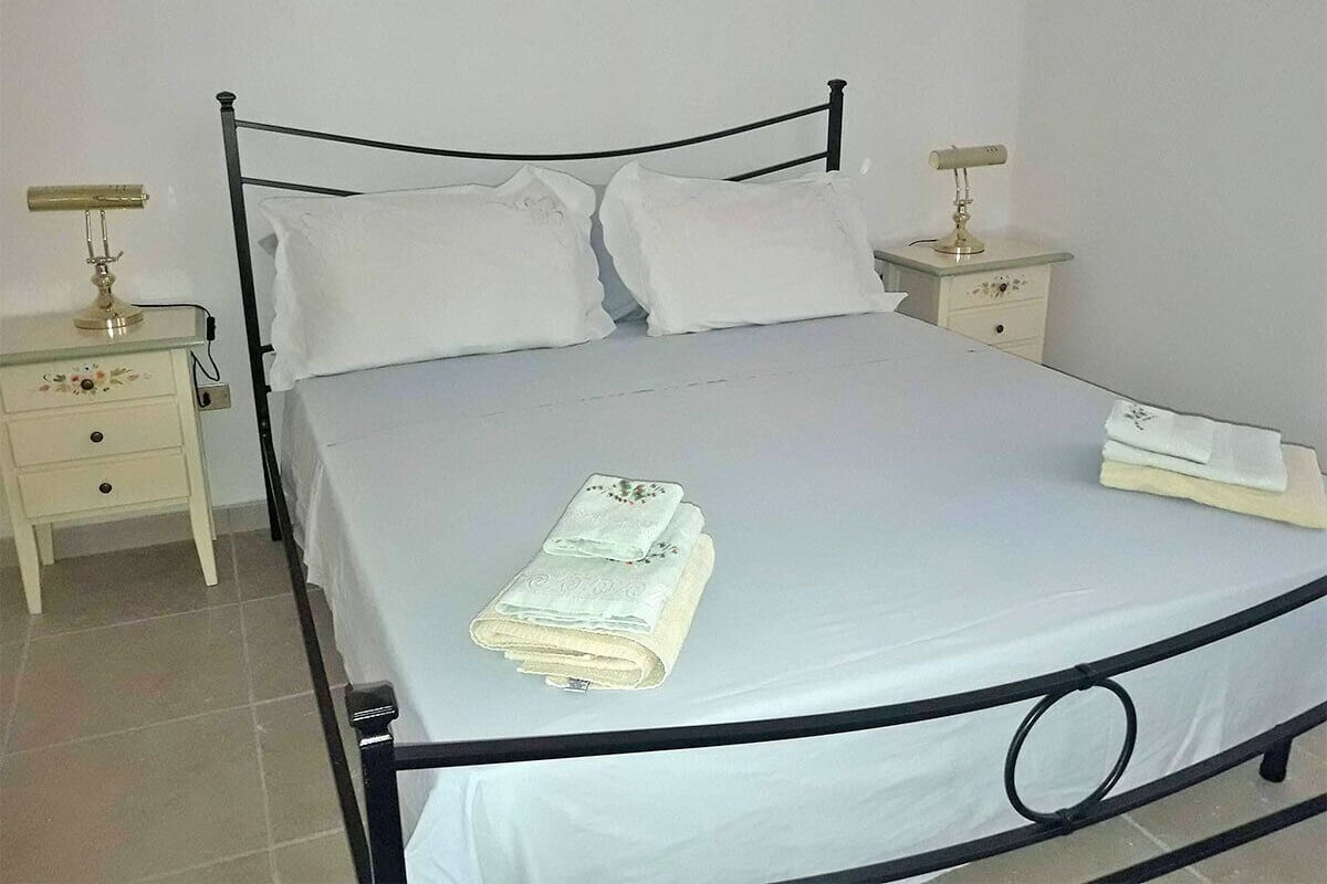 Un lit bien fait, avec du linge de lit blanc et des serviettes pliées, sur un cadre de lit en métal, dans une pièce aux murs blancs et au sol carrelé.
