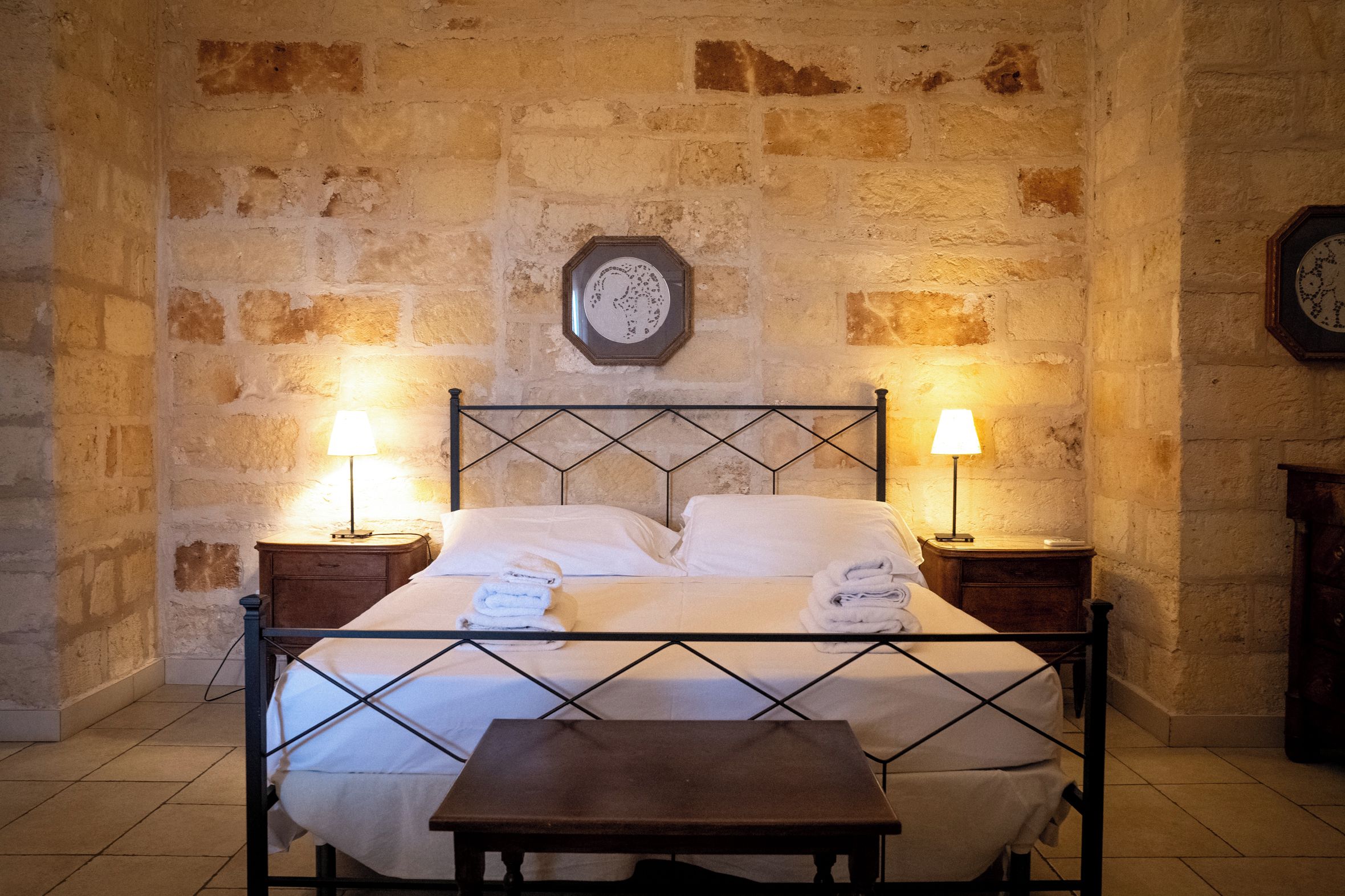 Una cama con estructura metálica, dos mesitas de noche con lámparas y un reloj de pared sobre la cama.