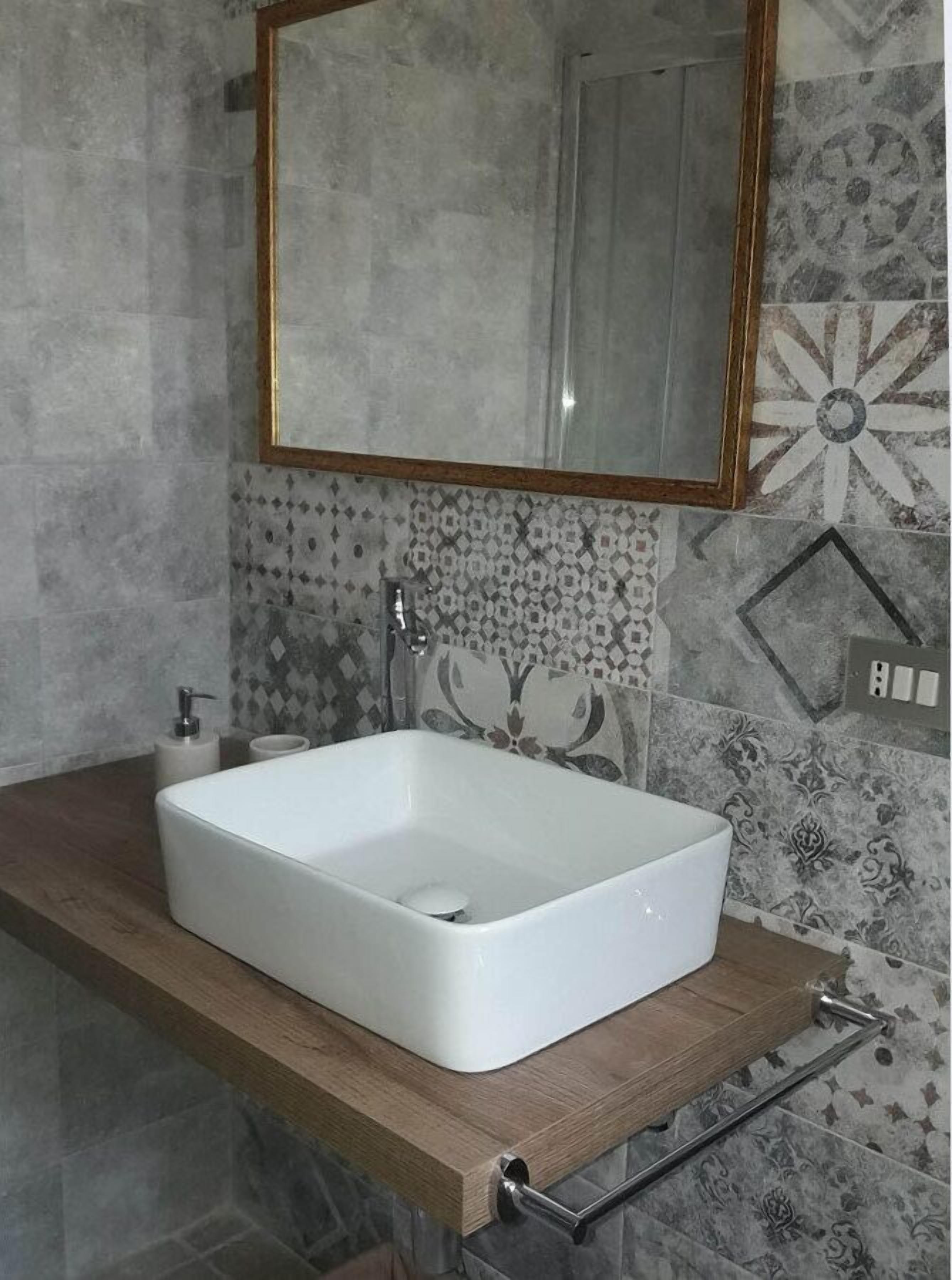 A bathroom with a white rectangular sink, a wooden countertop, a mirror with a gold frame, and patterned tiles on the walls.