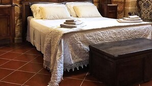 A bedroom with a stone wall, a wooden bed with a carved headboard, two bedside tables with lamps, a small bench, and a tiled floor. - Masseria Scianne (Nardò)