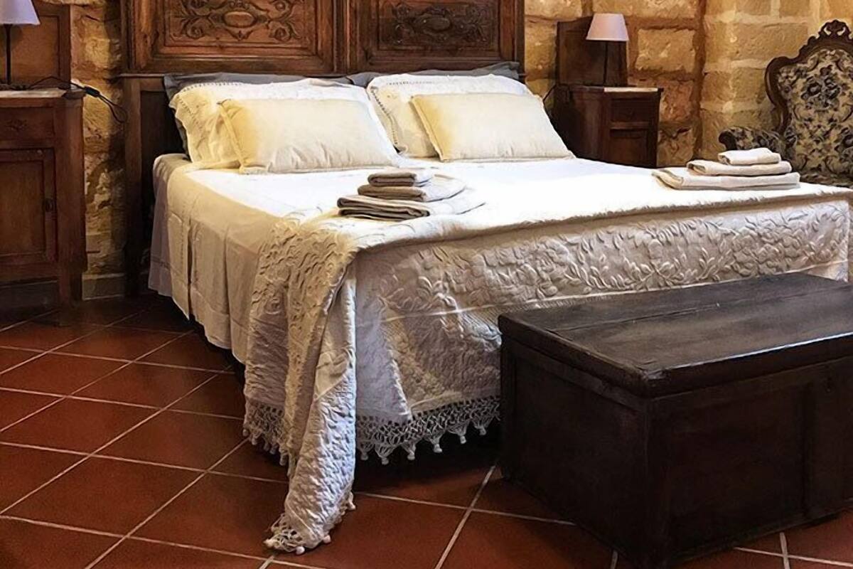 Une chambre avec un mur en pierre, un lit en bois doté d’une tête de lit sculptée, deux tables de chevet avec des lampes, un petit banc et un sol carrelé.
