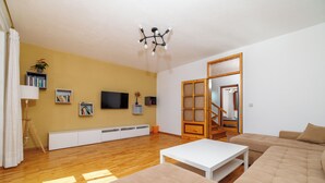 A living room with a wooden floor, a white sofa, a white coffee table, a TV mounted on the wall, and a bookshelf.