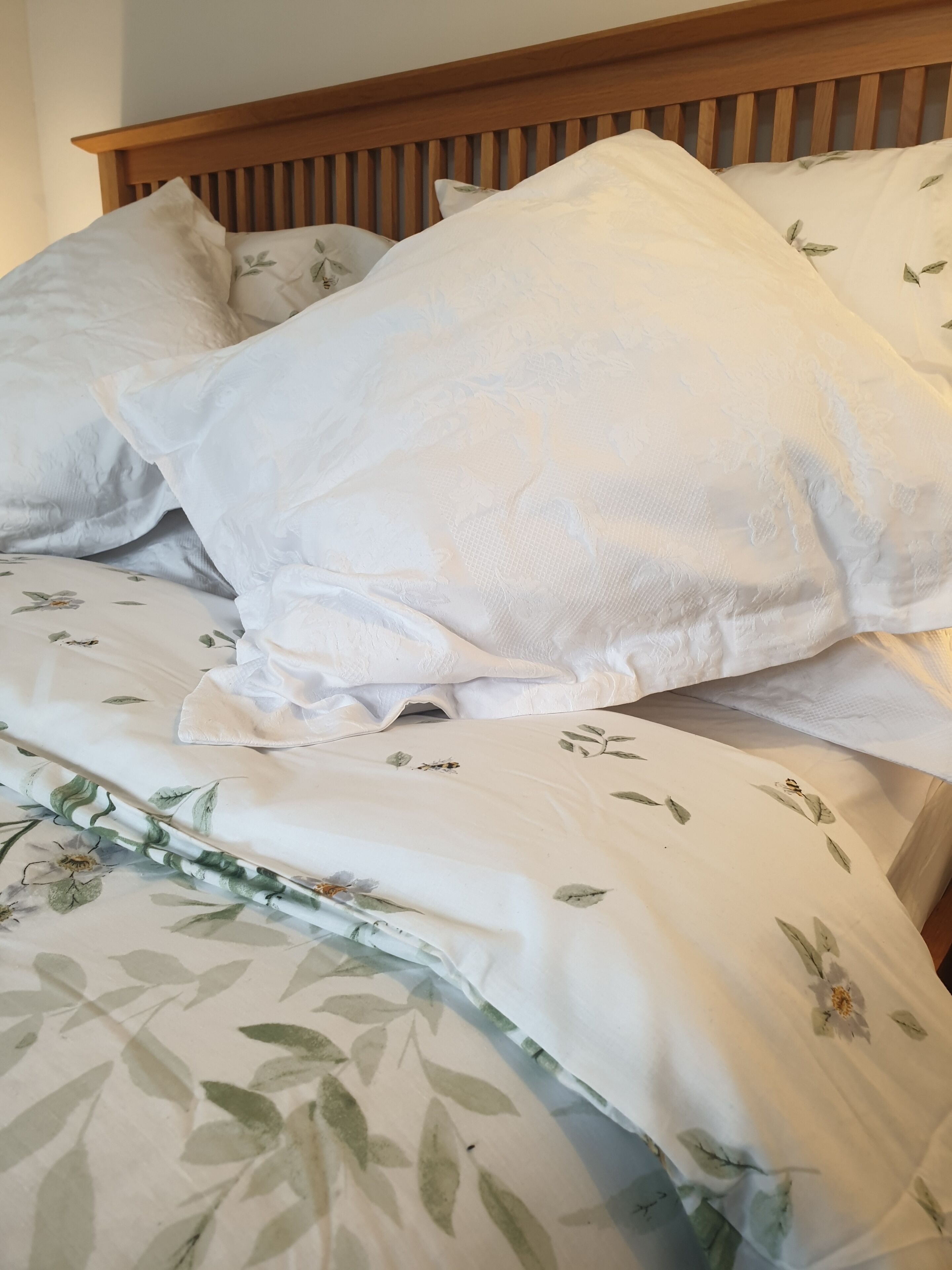 Una cama con ropa de cama blanca y estampados florales, cabecera de madera y varias almohadas.