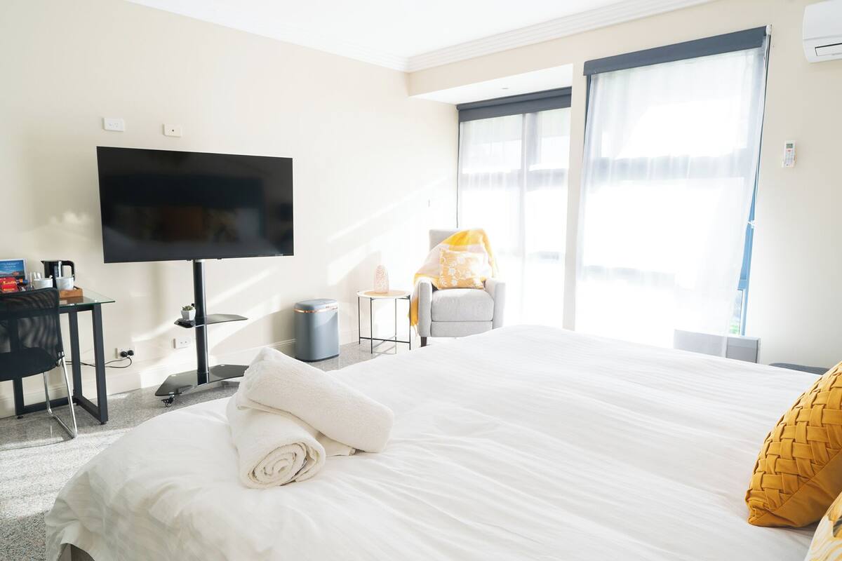 A hotel room with a bed, a TV, a chair, a small table, and a window with curtains.