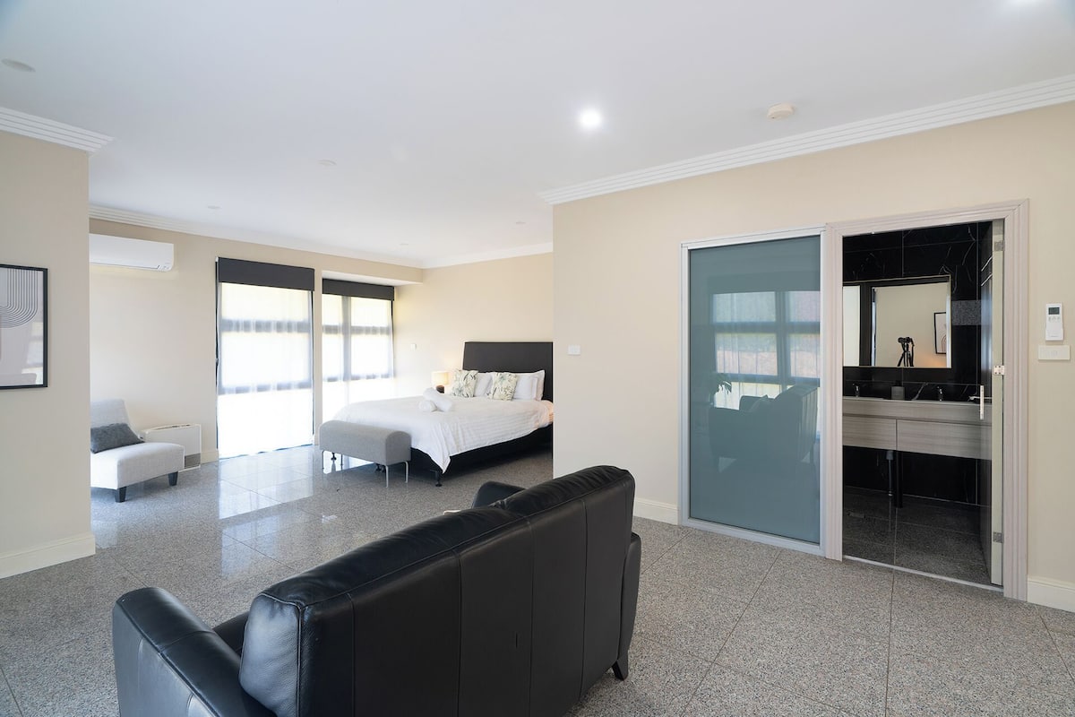 A modern hotel room with a large bed, a seating area, a bathroom with a mirror, and a sliding door to the outside.