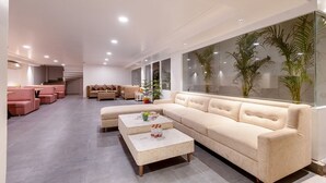 A modern living room with a beige sectional sofa, a coffee table, and a glass wall with plants. - The Pinky Villa Heritage Boutique Suites (New Delhi)