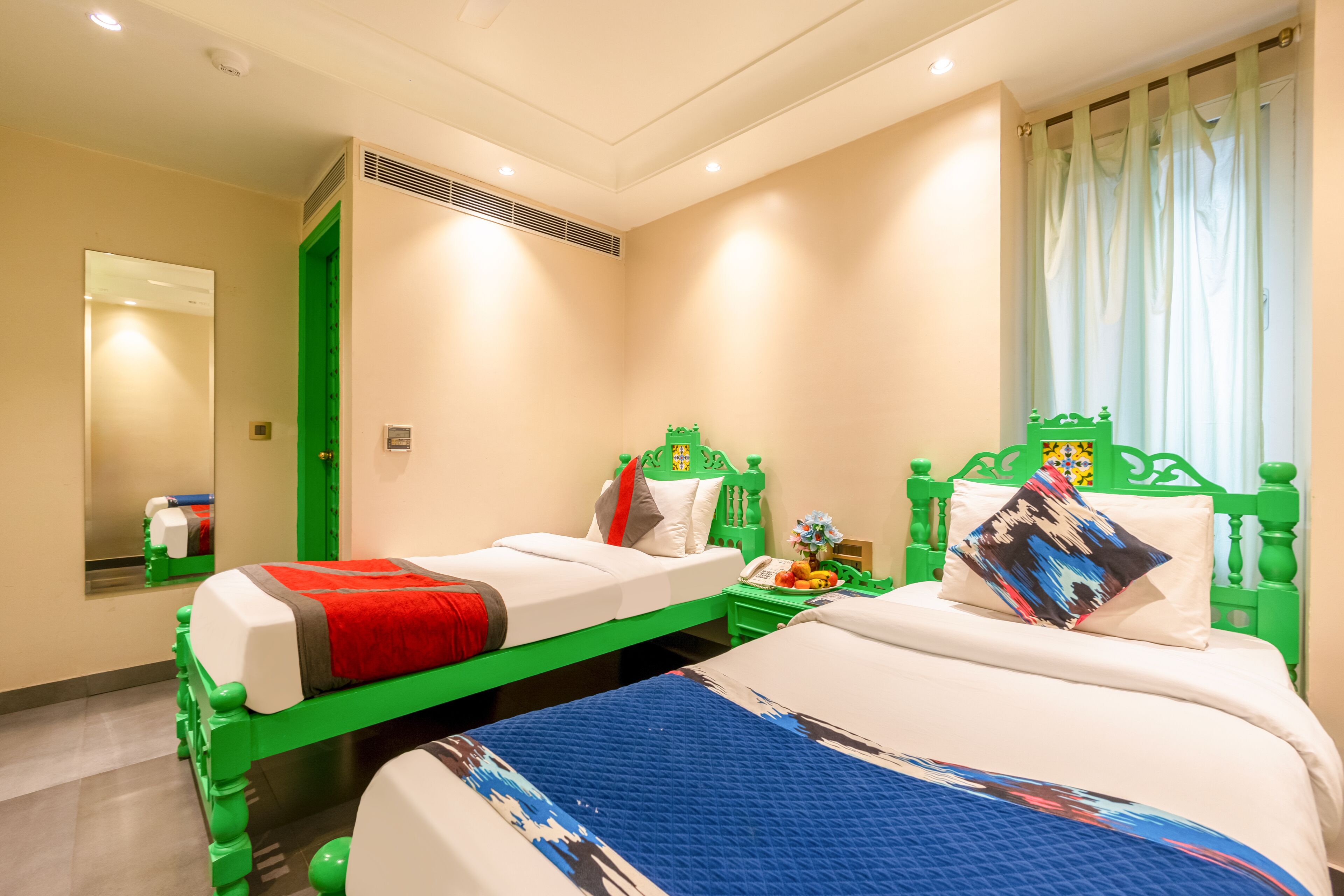 Een hotelkamer met twee bedden, een spiegel, een groen hoofdeinde en een raam met gordijnen.
