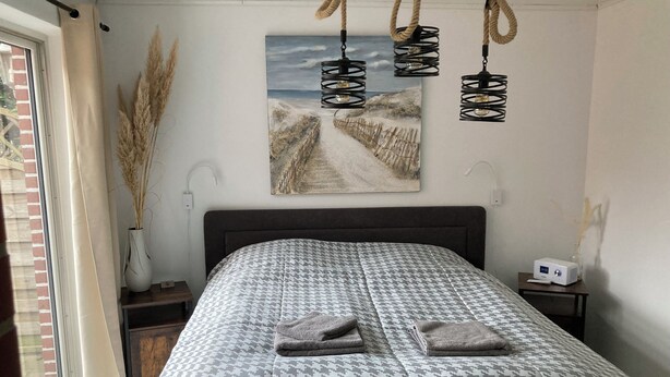 A bedroom with a patterned bedspread, a painting of a beach scene, and a bedside table with a lamp and a device. - Work & Relax - Your workation / vacation apartment on the North Sea with stable WLAN (Eddelak)