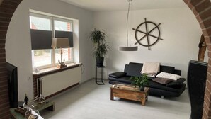 A living room with a black sofa, a wooden coffee table, a potted plant, a ship's wheel decoration, and a fireplace. - Work & Relax - Your workation / vacation apartment on the North Sea with stable WLAN (Eddelak)