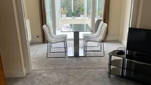 Une pièce dont le sol est recouvert d’une moquette, avec une table en verre et deux chaises, un téléviseur posé sur un meuble et une vue dégagée sur l’extérieur grâce à une grande fenêtre.
