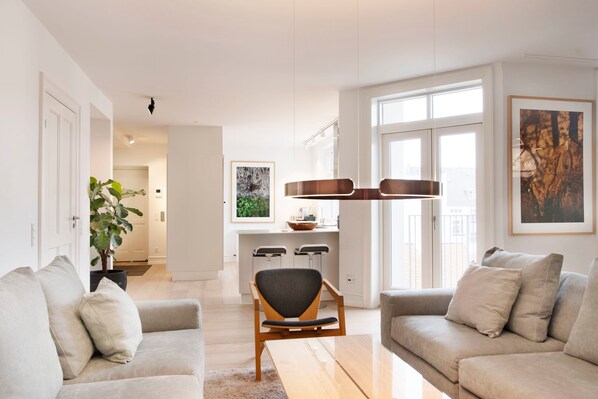 Un salon moderne avec un canapé, une table basse et un coin repas. On aperçoit une cuisine au fond, avec un tabouret de bar et une plante.