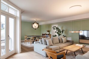 Un salon moderne avec un canapé d’angle, une table basse et un coin repas comprenant une table et des chaises. Une grande fenêtre offre une vue sur l’extérieur, et un téléviseur est fixé au mur.