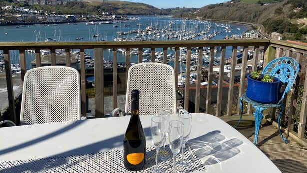 Un balcón con una mesa, sillas, una botella de vino y copas. Se aprecia una marina con barcos y una ciudad al fondo.