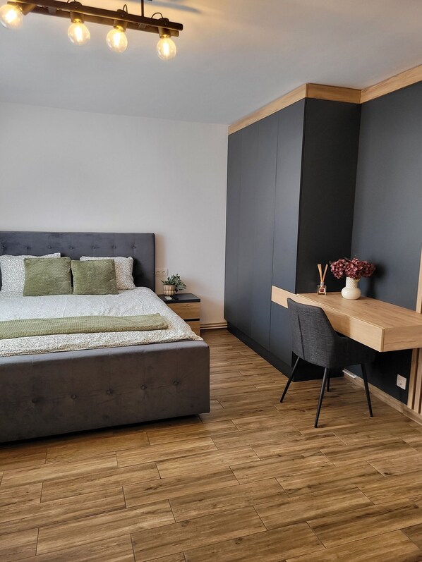 A bedroom with a large bed, a desk, and a chair. There is a nightstand with a plant and a vase. - Stella Cozy Apartament (Targu Mures)