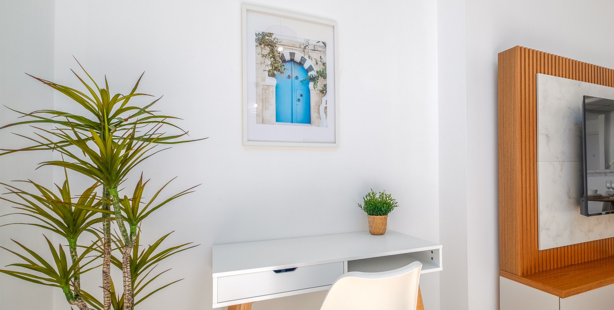 A modern room with a white desk, a framed picture on the wall, a small potted plant, and a wooden cabinet.