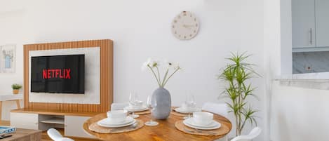 A dining area with a wooden table set for four, white chairs, a vase with flowers, and a wall-mounted clock.