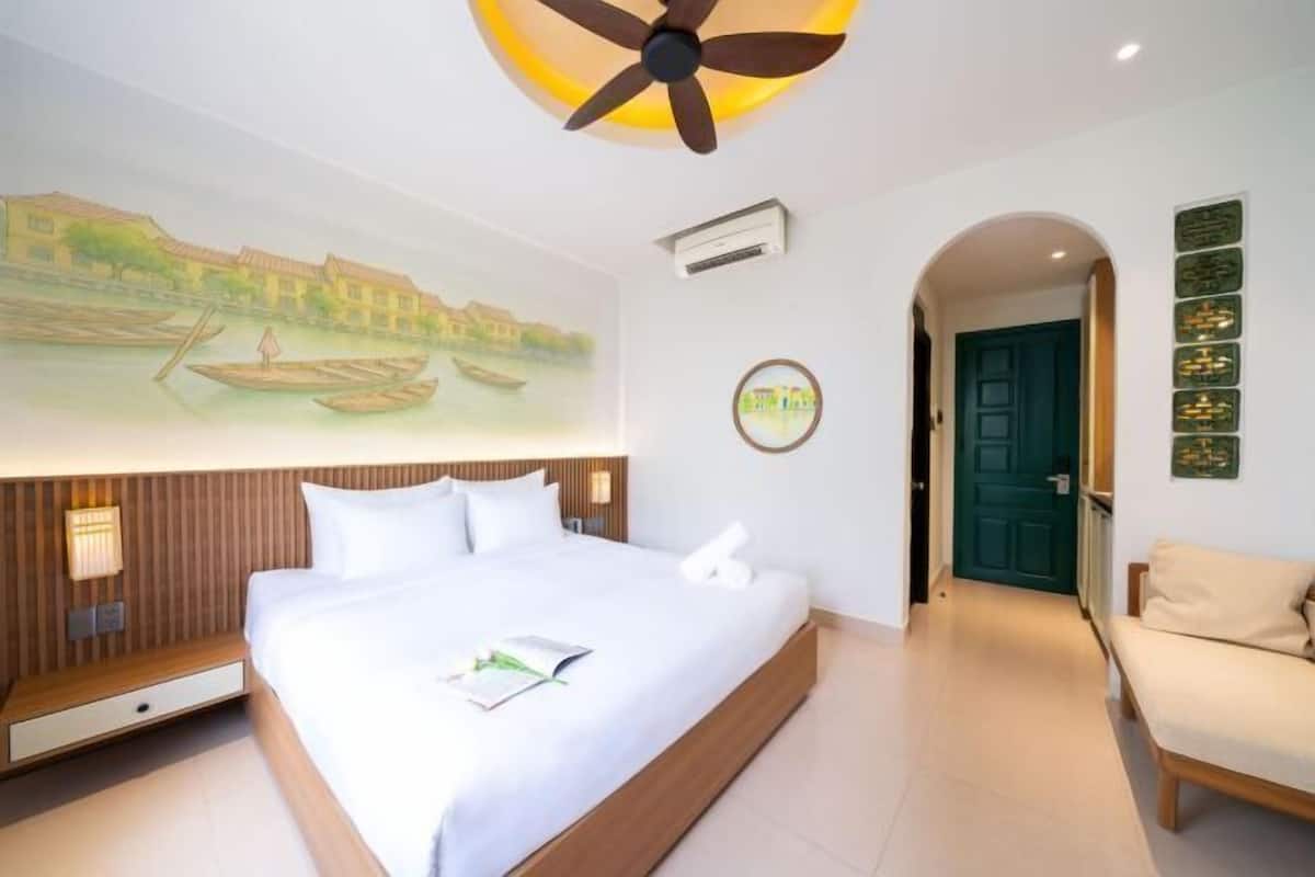 Camera d'albergo con un letto, una ventola a soffitto, un condizionatore d'aria a parete, un quadro raffigurante un paesaggio fluviale, una porta e un'area salotto.