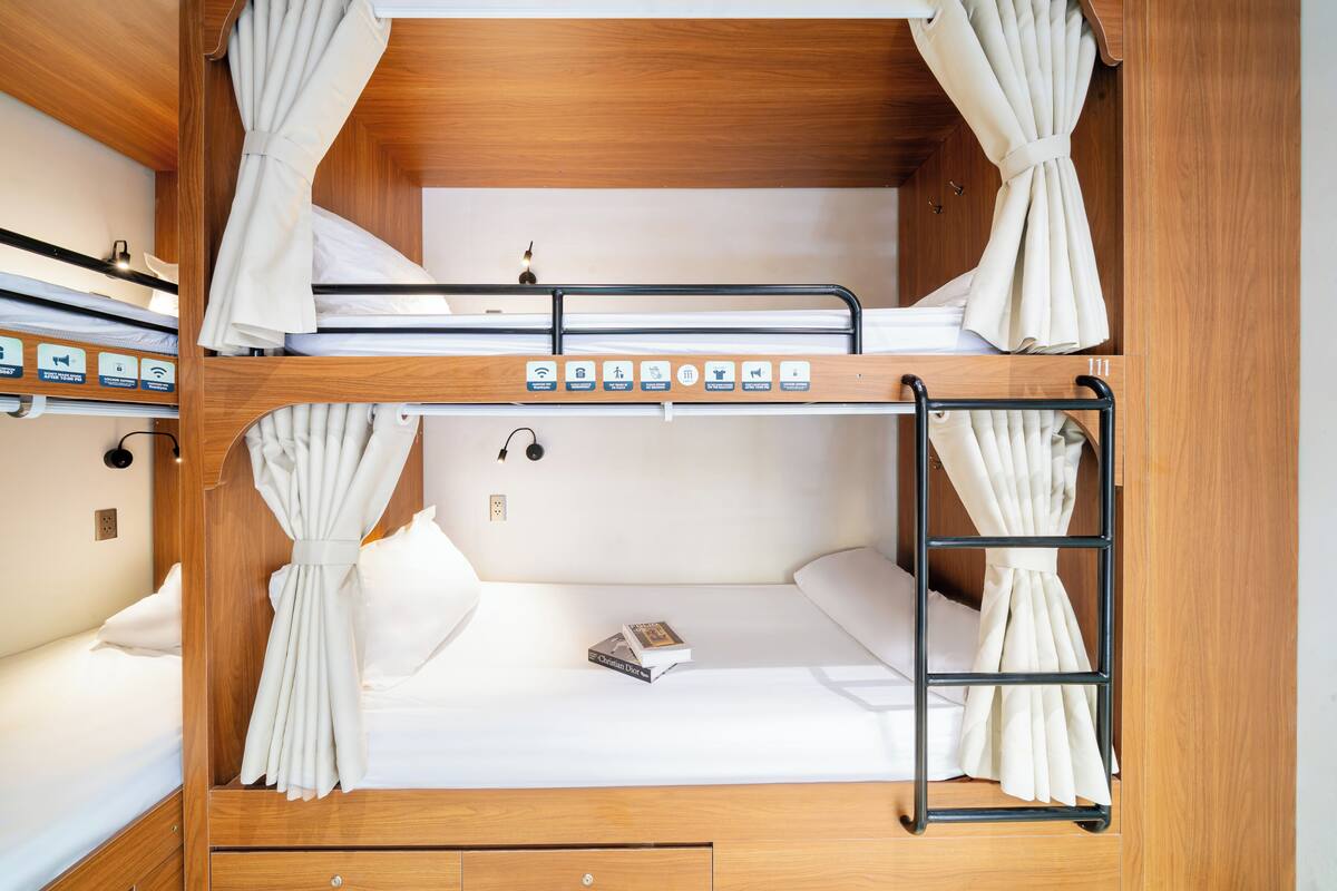 Letto a castello con struttura in legno, biancheria bianca e tende. Sono presenti pannelli di controllo sul letto e una scala applicata lateralmente.