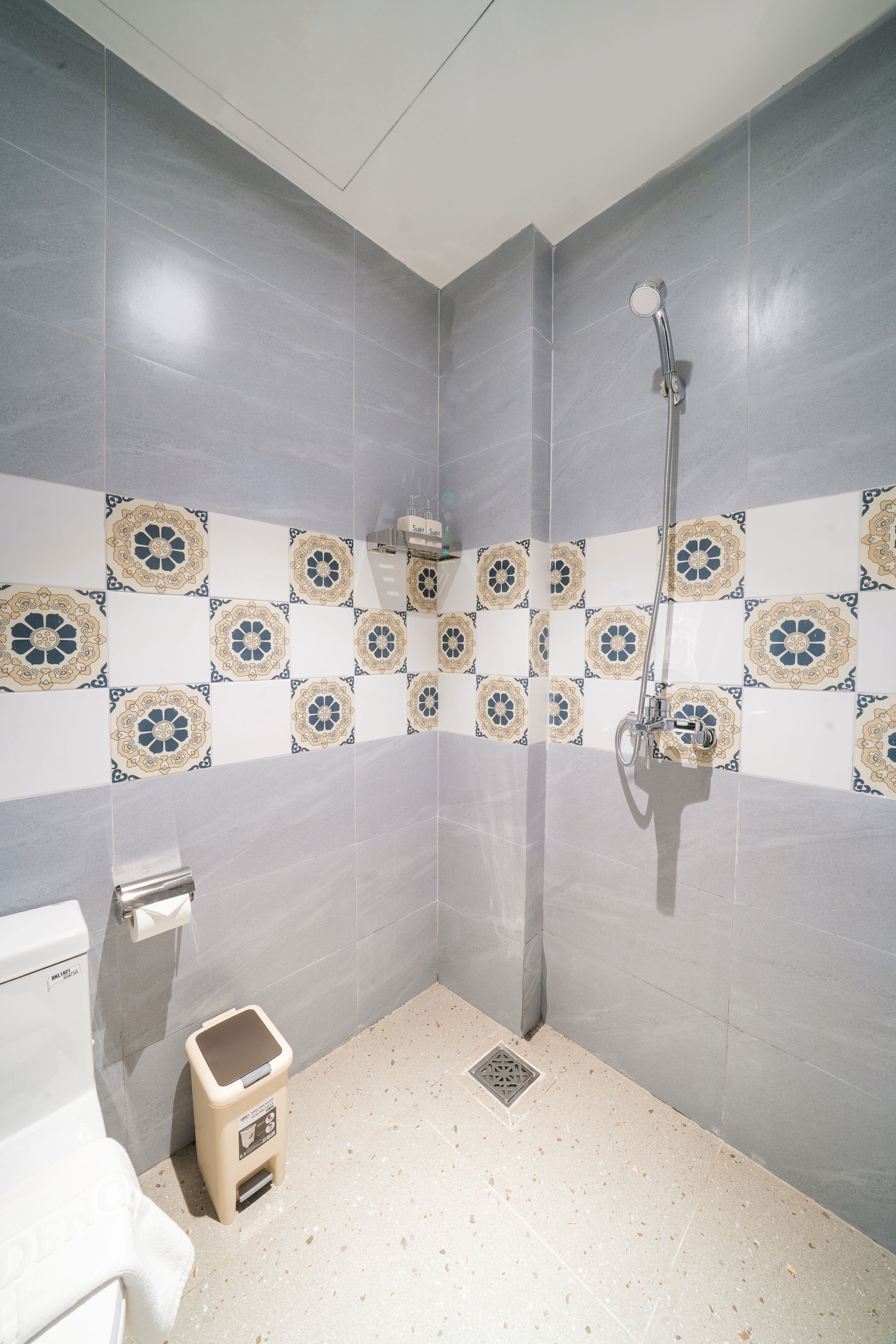 A bathroom with grey tiles, a shower, a toilet, and a trash bin.
