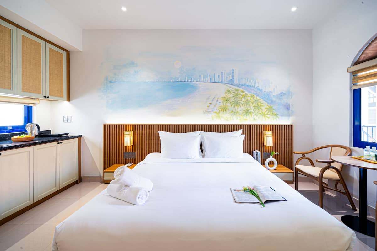 Camera d'albergo con un letto, un quadro raffigurante una spiaggia, un'area pranzo con tavolo e sedie e un angolo cottura.