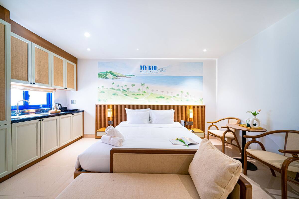A hotel room with a bed, a sofa, a small table with a vase, and a kitchen area with cabinets and a sink.