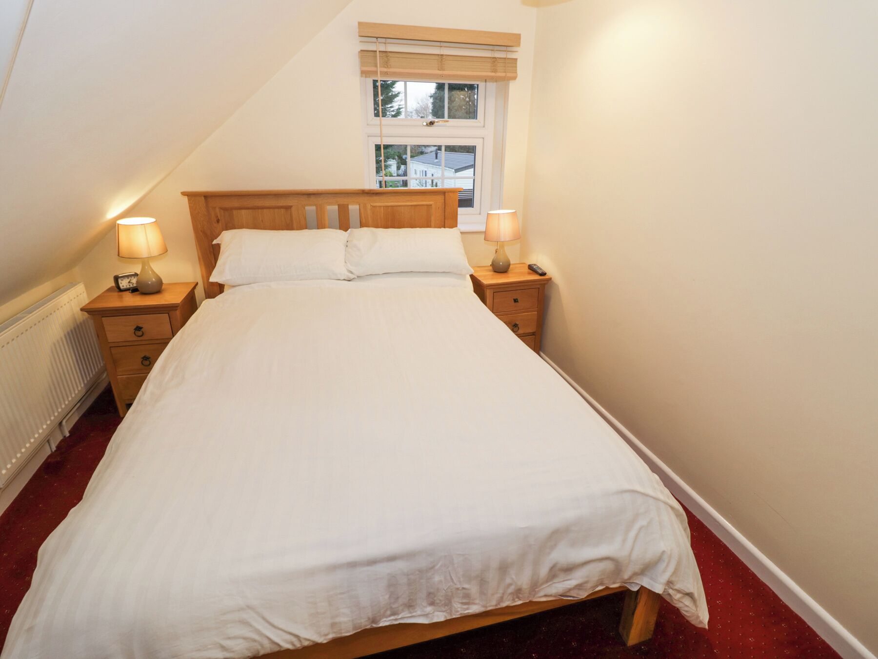A neatly made bed in a room with a sloped ceiling, two bedside tables with lamps, a window with blinds, and a radiator.