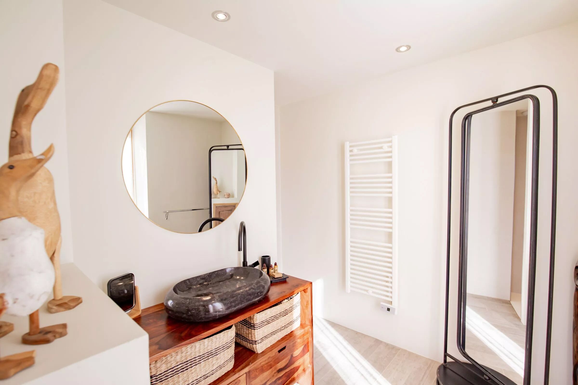 A modern bathroom with a round mirror, a wooden vanity, a large sink, and a tall mirror.