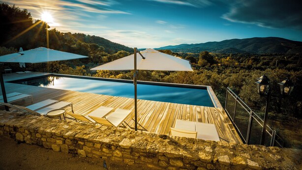 A swimming pool with wooden deck, lounge chairs, and umbrellas, surrounded by stone walls and overlooking a mountainous landscape. - Château les Gardis (Lourmarin)