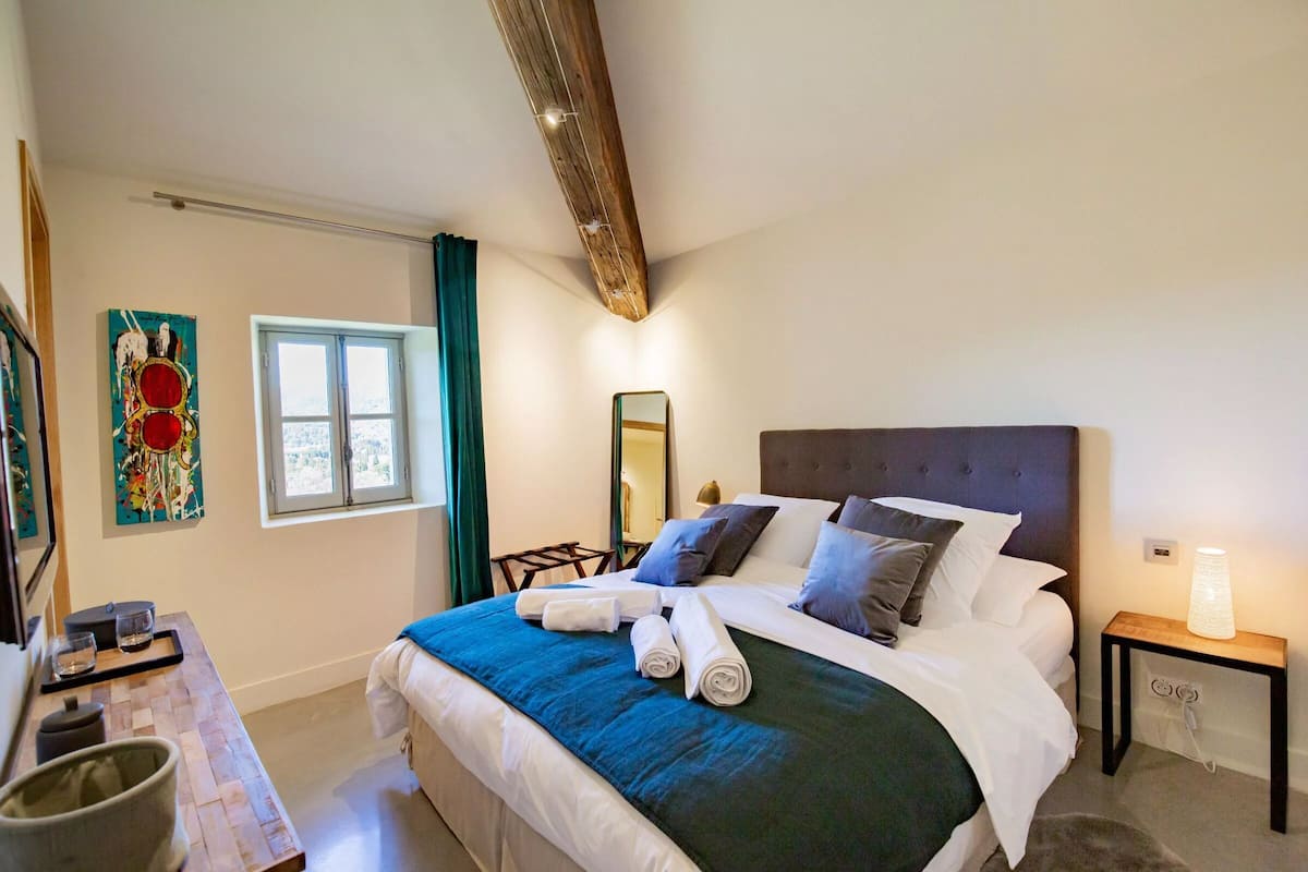 Een opgemaakt bed met kussens, een nachtkastje met een lamp, een raam met gordijnen en een wanddecoratie.