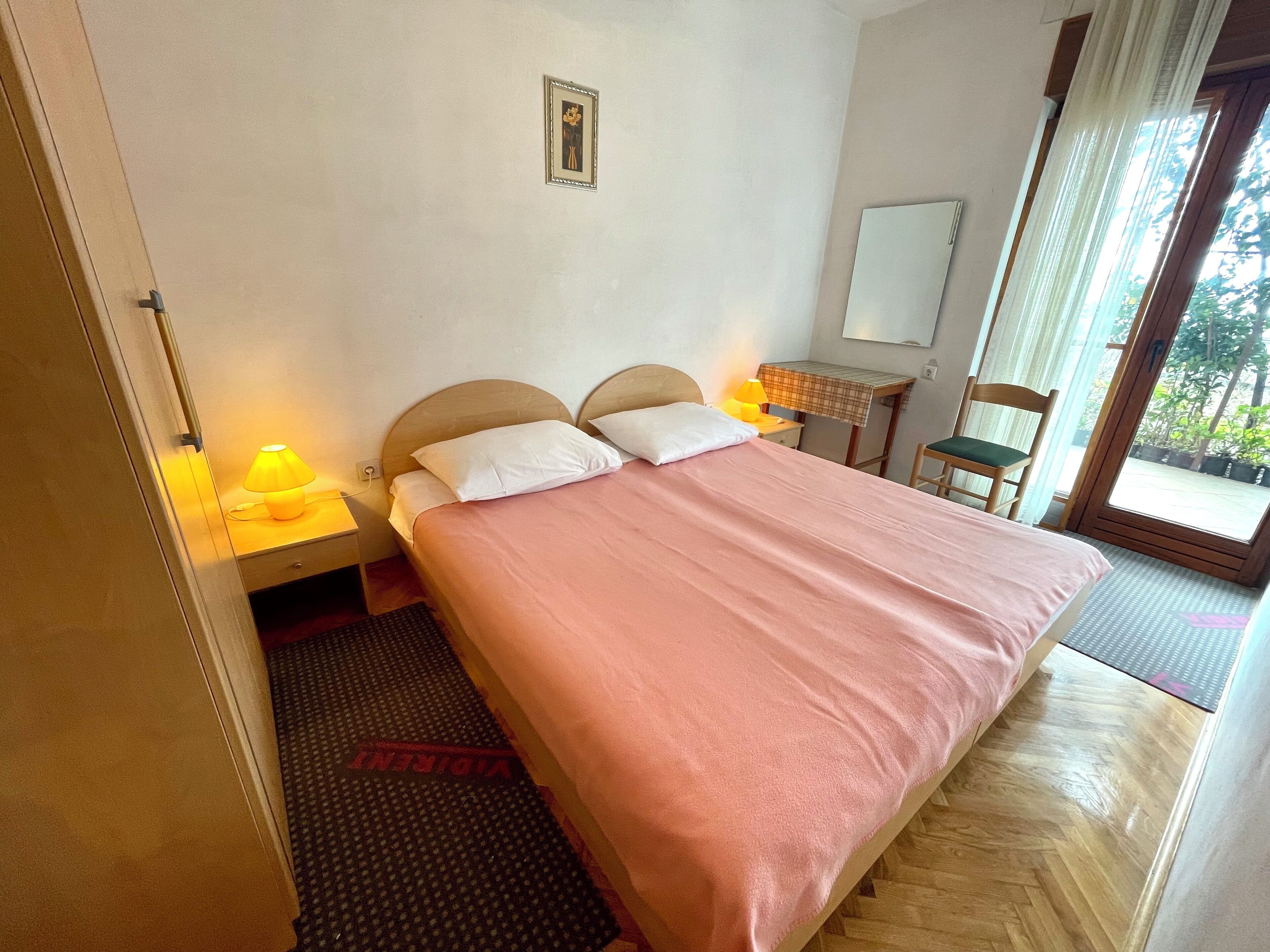 Une chambre d’hôtel avec un lit, des lampes de chevet, une petite table, une chaise, un miroir et une fenêtre donnant sur un paysage verdoyant.