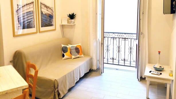 A small, well-lit room with a bed, a table, and a chair. There is a door leading to a balcony with a metal railing. Two framed pictures are on the wall above the bed. - Summer House (Trapani)