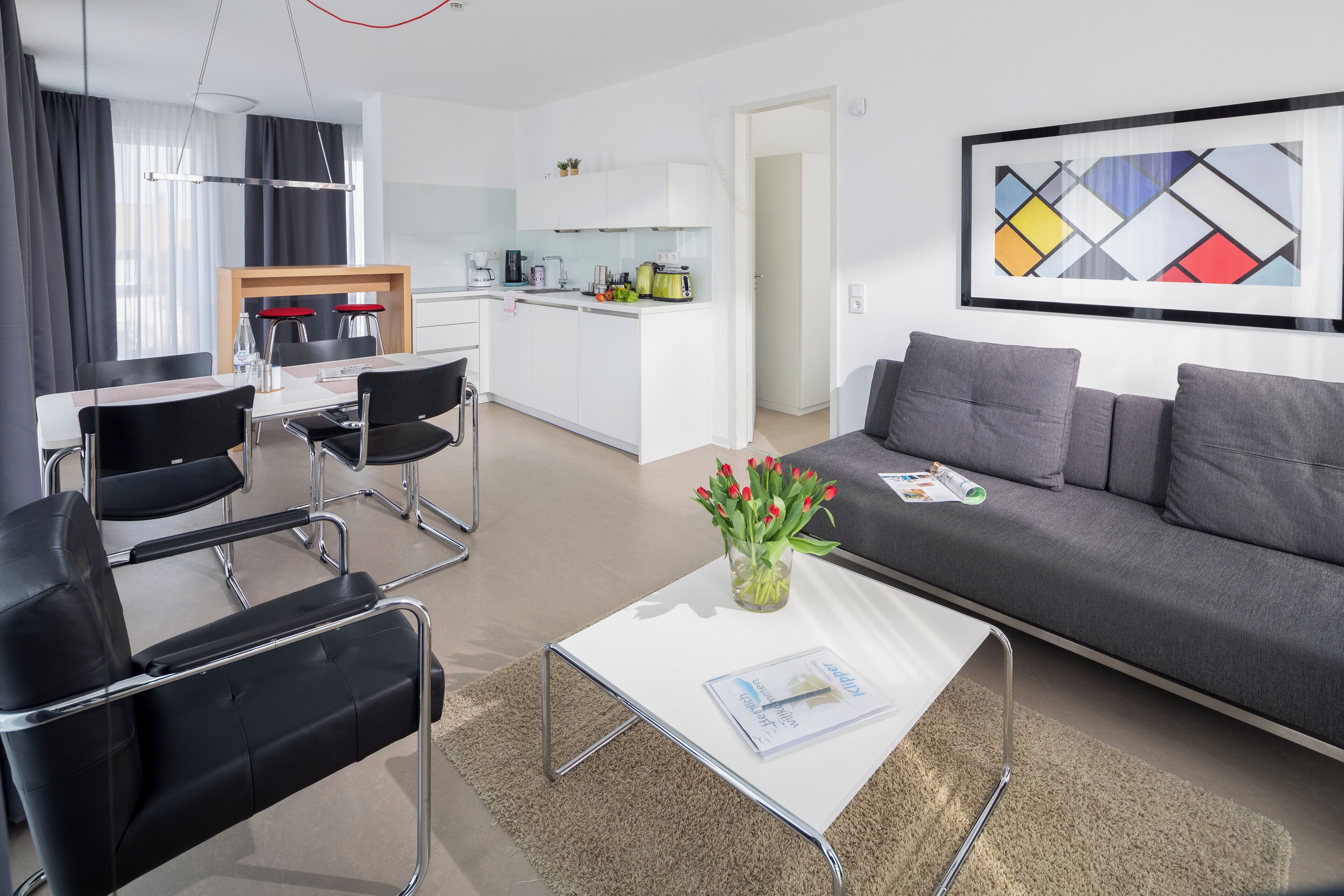 A modern living room with a grey sofa, black chairs, a white coffee table, and a kitchen in the background.