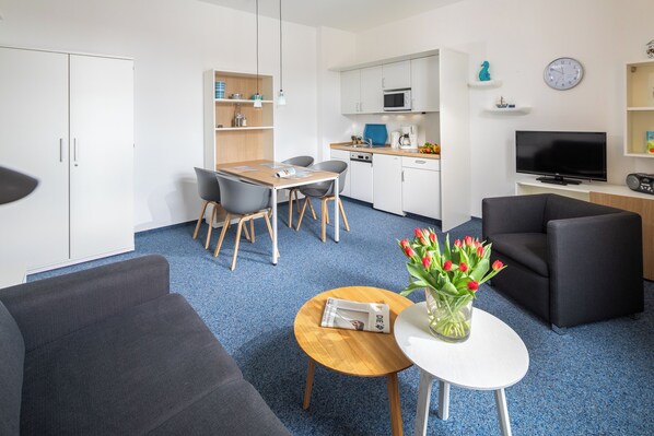 Una sala de estar moderna con un sofá, una mesa de comedor con sillas, una zona de cocina con microondas y cafetera, un televisor y un jarrón con flores.