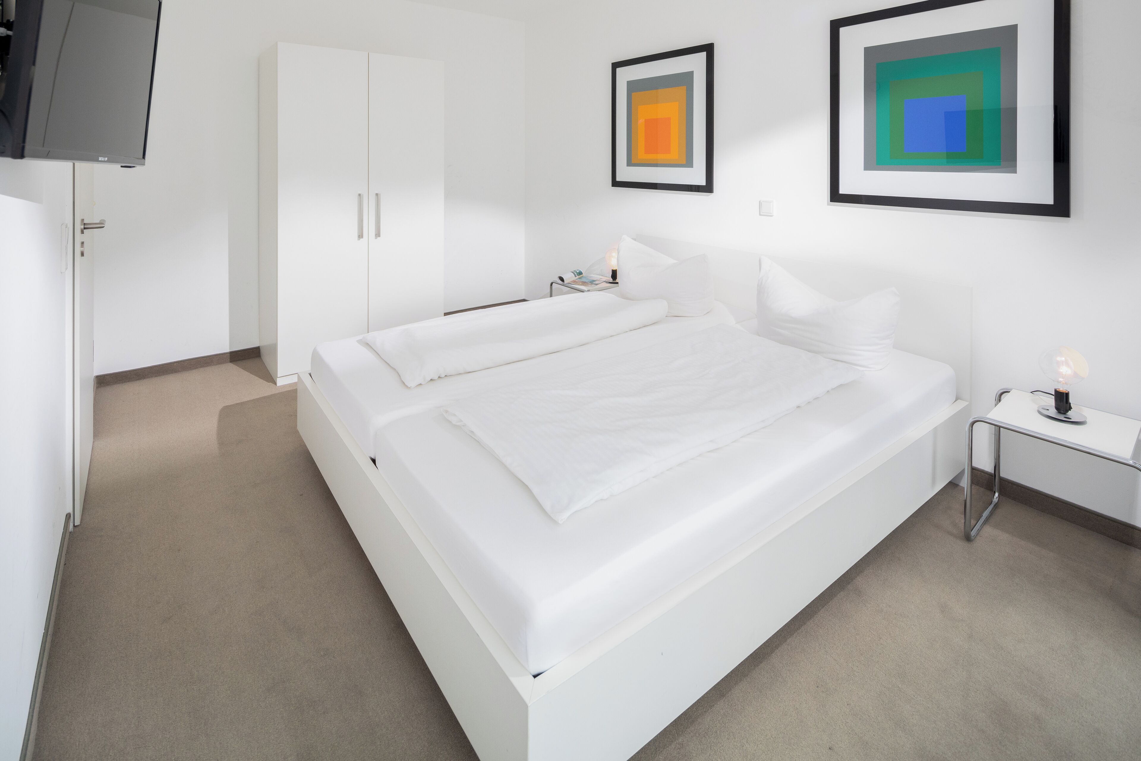 A modern hotel room with a large bed, a bedside table, a TV, and artwork on the wall.