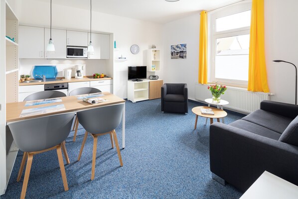 A modern living room with a sofa, a dining area with a table and chairs, a kitchen with white cabinets and appliances, and a window with yellow curtains. - Haus Klipper Norderney (Norderney)