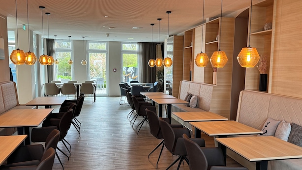 Interior moderno de restaurante com mesas e cadeiras de madeira, iluminação pendente e janelas amplas.