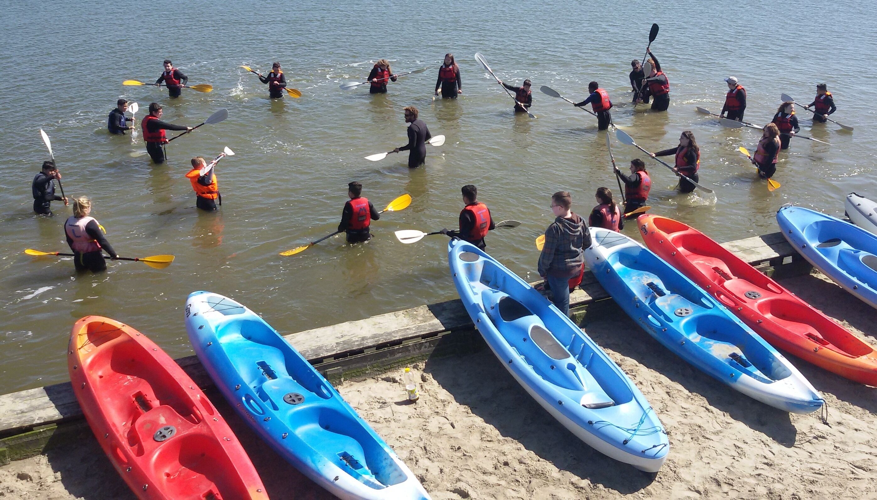 A group of people in kayaks and life jackets on a sandy shore, preparing to paddle.