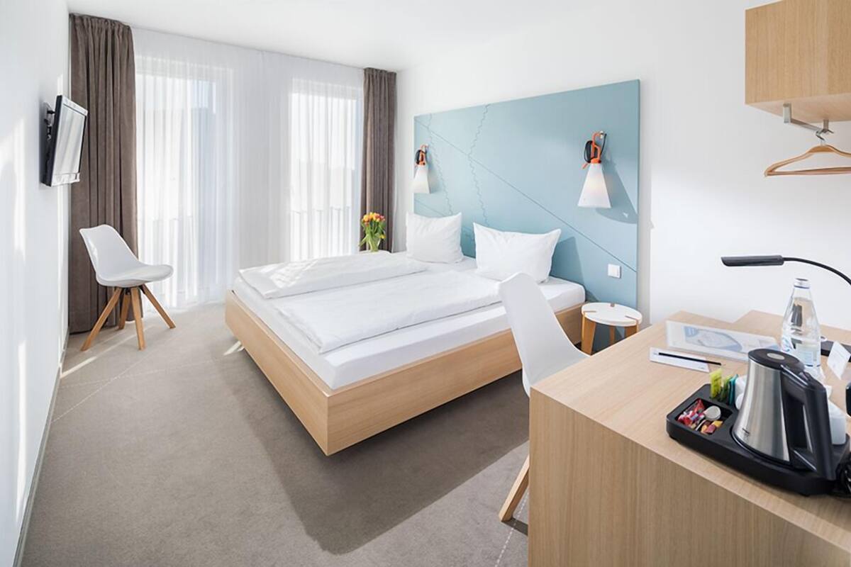 Ein modernes Hotelzimmer mit einem großen Bett, einem Schreibtisch mit Kaffeemaschine, einem Sessel und einem an der Wand montierten Fernseher.