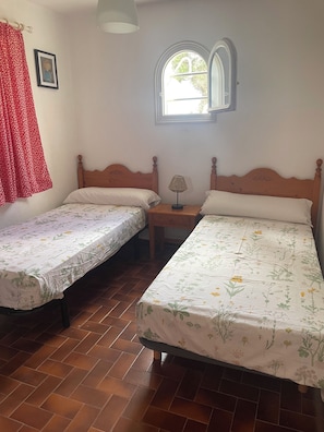 A room with two single beds, a small table with a lamp, a window with a view, and a red curtain. - House with pool near the beach and marina (Cala en Bosc)