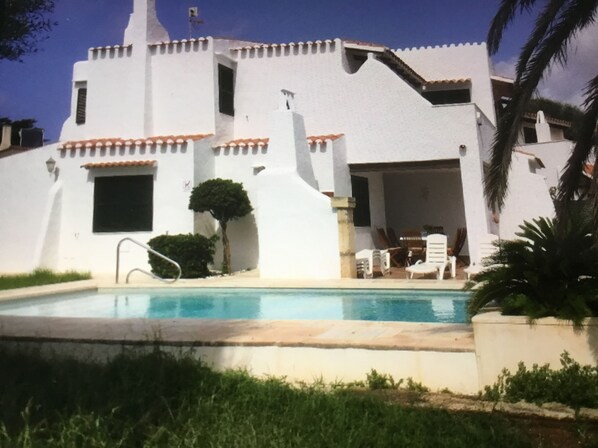 Un bâtiment blanc avec une piscine, une terrasse avec des chaises et un petit arbre.