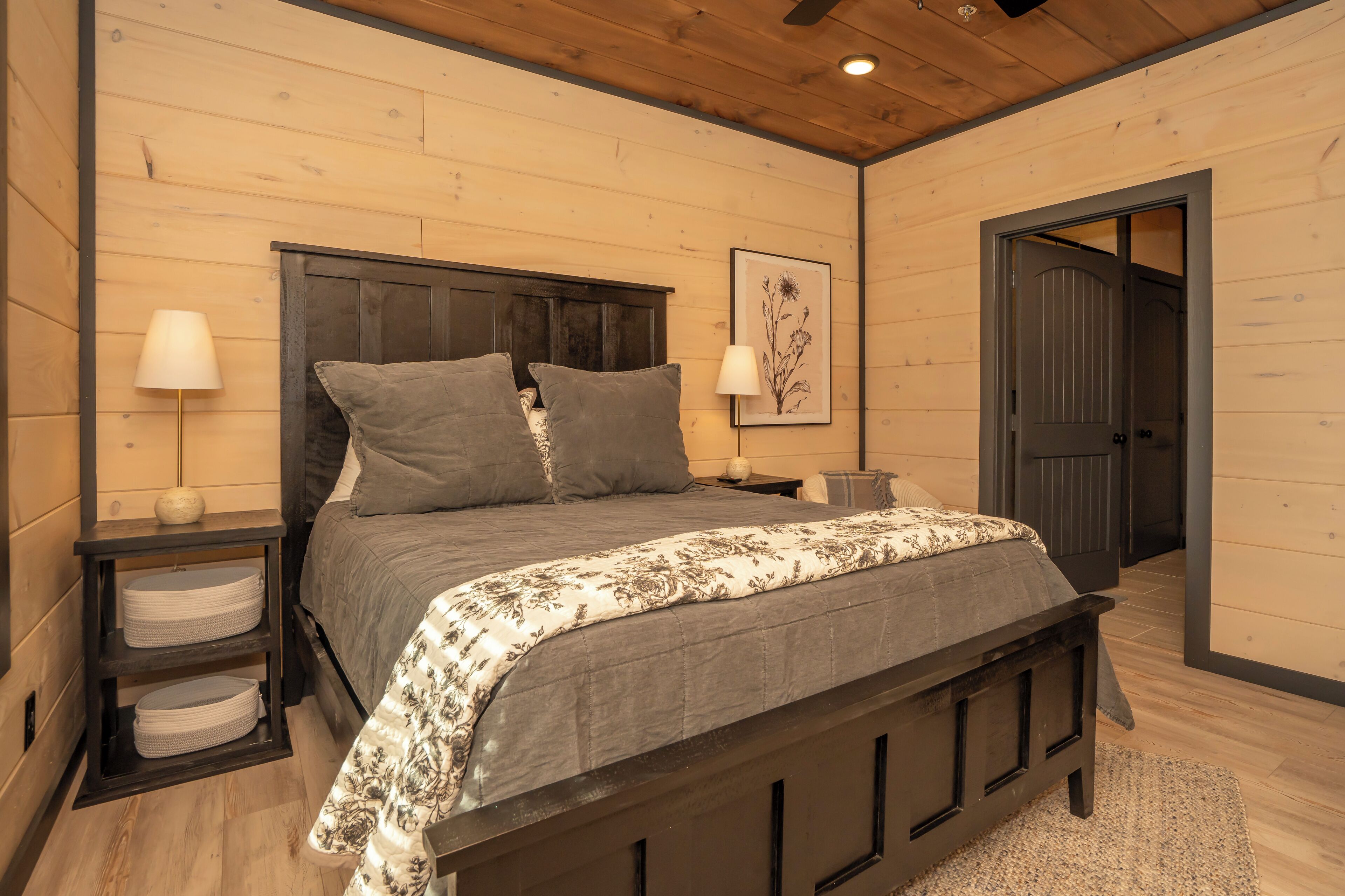 A bedroom with a wooden headboard, two bedside tables, a bed with grey bedding, a framed floral artwork, and a door leading to another room.