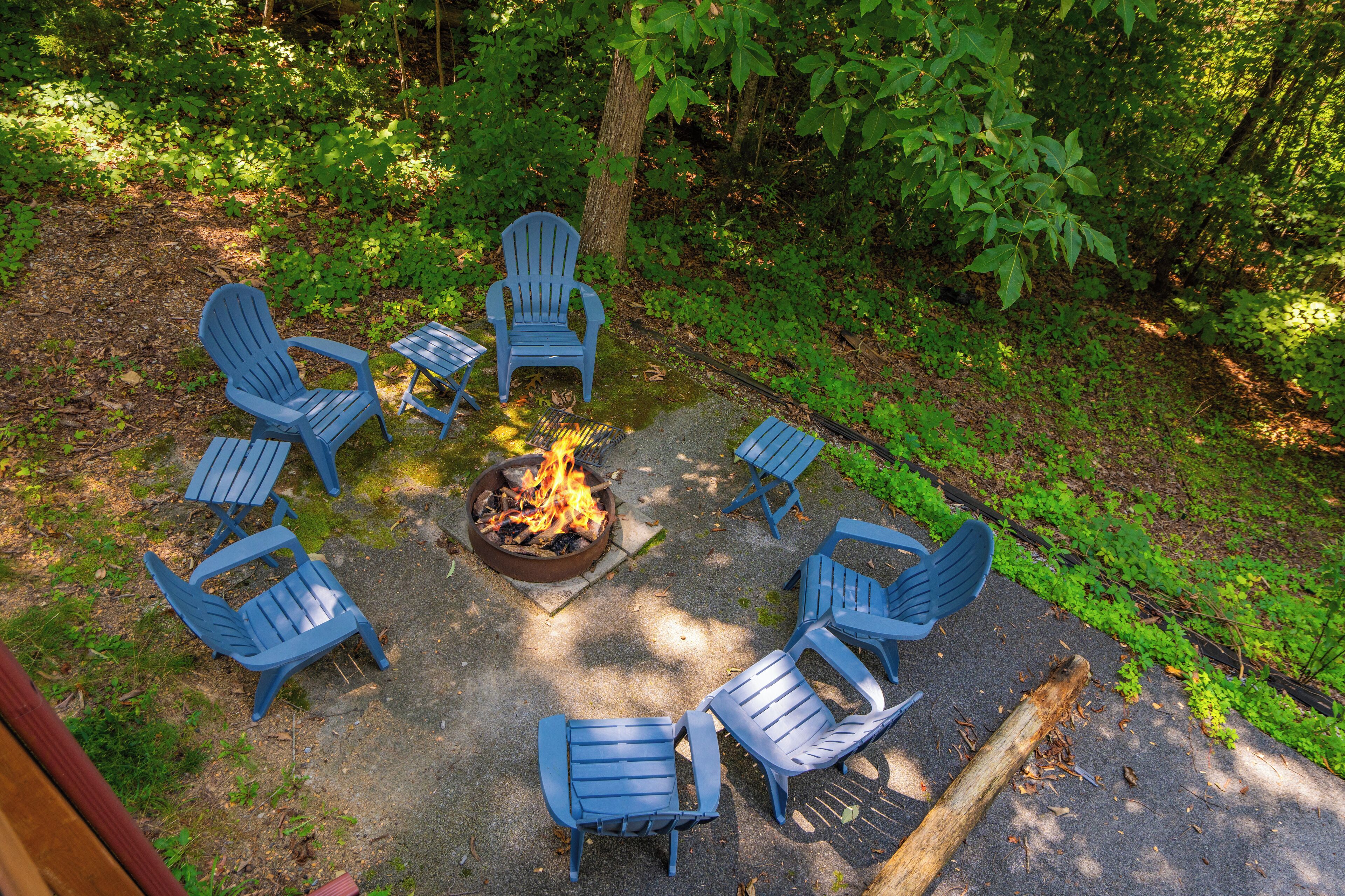 A circular arrangement of blue Adirondack chairs around a central fire pit in a wooded area.