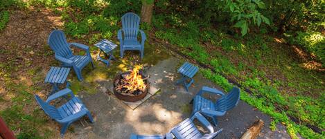 A circular arrangement of blue Adirondack chairs around a central fire pit in a wooded area.
