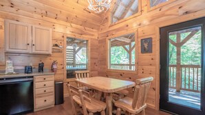 Interior de una cabaña de madera con una mesa de comedor y sillas, una zona de cocina y ventanas amplias con vistas al bosque.