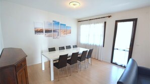 A dining area with a white table and black chairs, a wooden cabinet, and a large window with curtains. - Charming House With Private Fenced Garden in Lignano Pineta (Lignano Sabbiadoro)