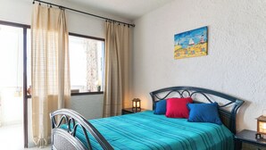 A bedroom with a bed, curtains, a nightstand, a wall painting, and a view through a window.
