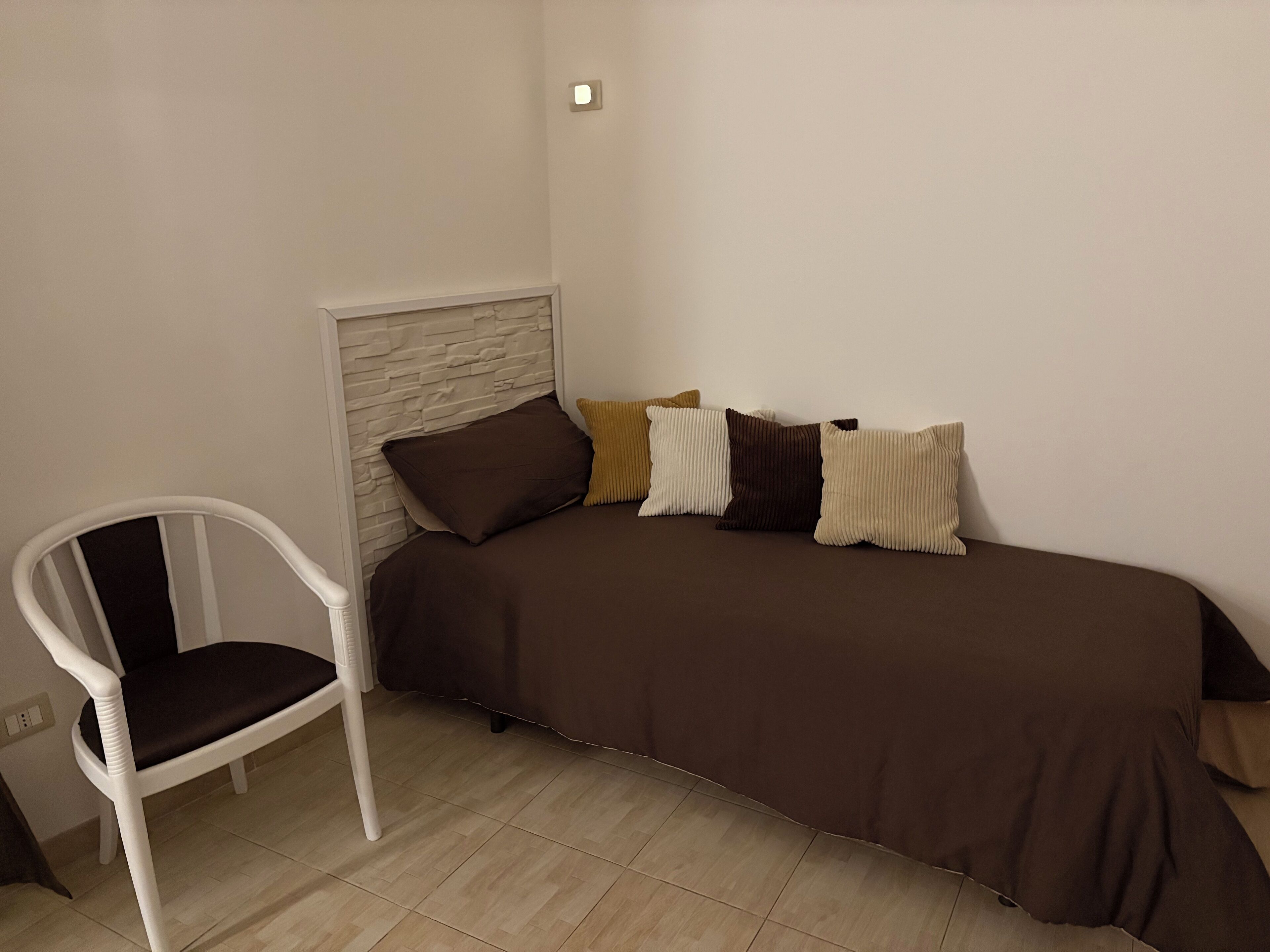 Ein kleines, ordentlich eingerichtetes Schlafzimmer mit einem Einzelbett, einem weißen Stuhl und einem weißen Kopfteil.