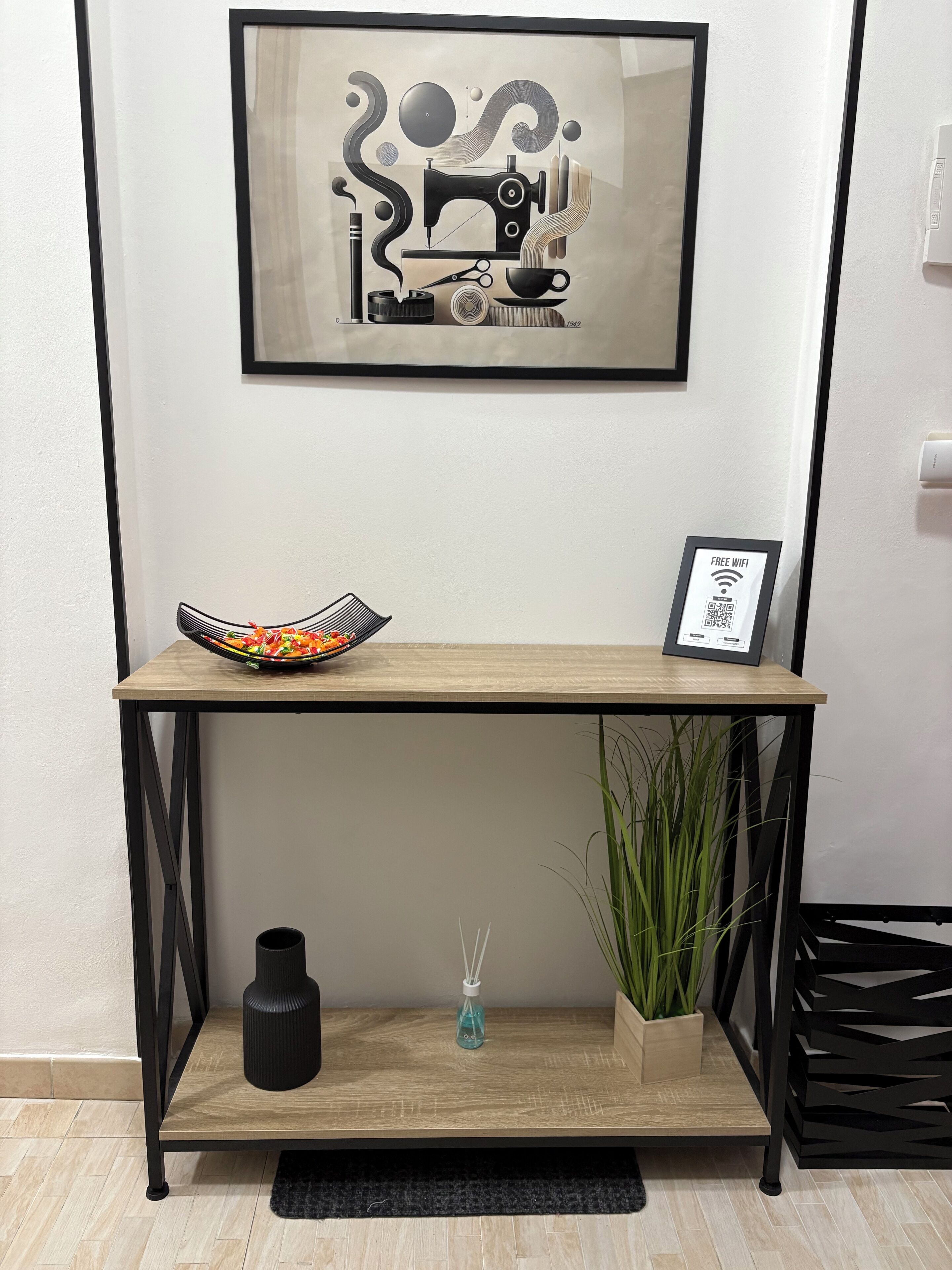 A framed abstract artwork on a wall, a wooden console table with a bowl of fruit, a framed picture, a black vase, and a potted plant.